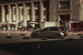 A sleek, dark-colored car is moving swiftly through an urban setting, surrounded by traffic cones and tires, suggesting a driving course or training area. The background features a large building with columns and several shaded awnings, contributing to a sense of speed and motion blur.