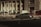 A sleek, dark-colored car is moving swiftly through an urban setting, surrounded by traffic cones and tires, suggesting a driving course or training area. The background features a large building with columns and several shaded awnings, contributing to a sense of speed and motion blur.