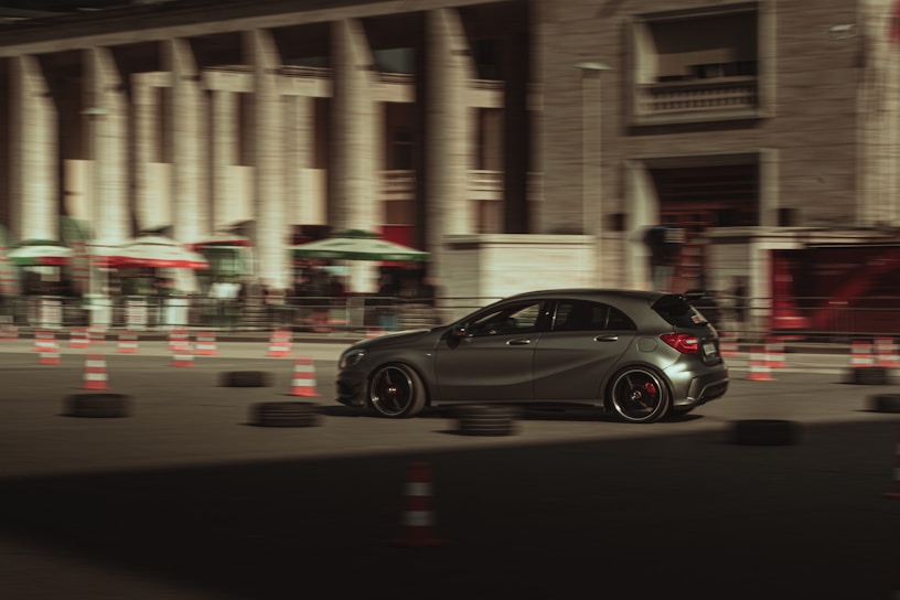 A sleek, dark-colored car is moving swiftly through an urban setting, surrounded by traffic cones and tires, suggesting a driving course or training area. The background features a large building with columns and several shaded awnings, contributing to a sense of speed and motion blur.