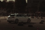A white car is navigating through a makeshift course outlined by traffic cones and tire barriers. The setting is outdoors, likely an automotive event or car show, with a crowd of spectators in the background. The lighting is dim, creating a moody, dramatic atmosphere.