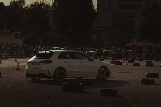 A white car is navigating through a makeshift course outlined by traffic cones and tire barriers. The setting is outdoors, likely an automotive event or car show, with a crowd of spectators in the background. The lighting is dim, creating a moody, dramatic atmosphere.