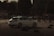 A white car is navigating through a makeshift course outlined by traffic cones and tire barriers. The setting is outdoors, likely an automotive event or car show, with a crowd of spectators in the background. The lighting is dim, creating a moody, dramatic atmosphere.