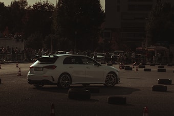A white car is navigating through a makeshift course outlined by traffic cones and tire barriers. The setting is outdoors, likely an automotive event or car show, with a crowd of spectators in the background. The lighting is dim, creating a moody, dramatic atmosphere.
