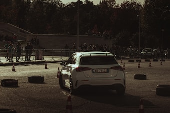 A white car is maneuvering through a course marked by traffic cones and tires. The scene is set in an open paved area with a crowd of spectators standing behind a metal barrier. The lighting appears to be subdued, possibly during late afternoon or early evening, casting shadows across the ground.