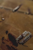 A razor blade lies on a surface that appears to be stained with a dark, reddish-brown substance. The background is blurred, making the razor blade and stains the focal points of the image.
