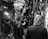 A bustling market scene in black and white with several people walking and interacting. The market is filled with shops selling various goods, including leather belts and woven fabrics. The overhead shelter and the crowd create a lively atmosphere.