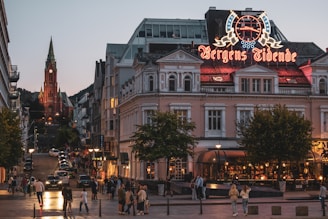 A lively urban street scene during dusk with a large building prominently displaying a neon sign. Many people walk on the street, and several cars are parked along the sides. The street leads up to a striking red brick church with a tall spire in the background.