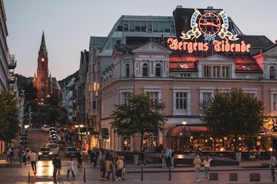 A lively urban street scene during dusk with a large building prominently displaying a neon sign. Many people walk on the street, and several cars are parked along the sides. The street leads up to a striking red brick church with a tall spire in the background.