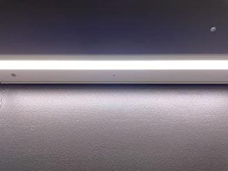 A minimalistic and modern light fixture is mounted on a smooth, dark surface. The lighting strip provides a bright, even illumination along its entire length, casting a subtle glow onto the surface below.
