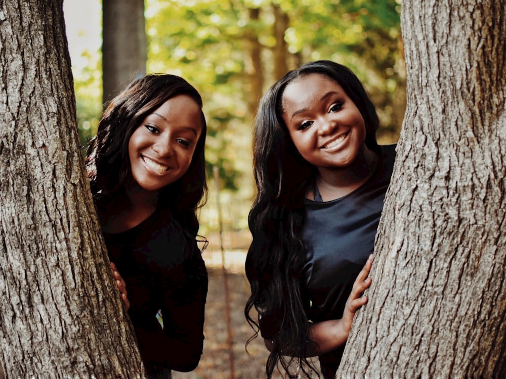 two women standing next to each other behind a tree