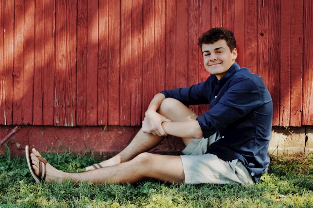 a young man sitting on the grass in front of a red barn