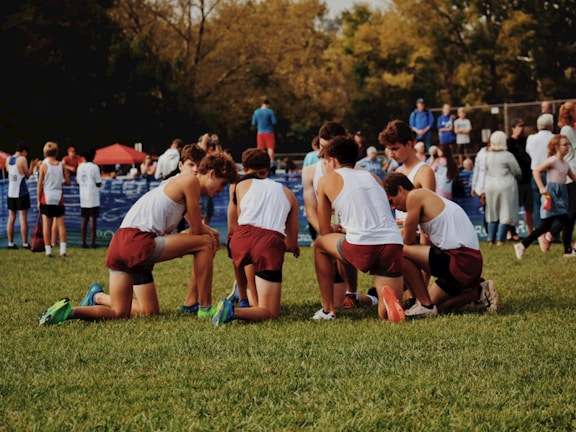 A group of young athletes wearing maroon shorts and white shirts gather on a grassy field, preparing for an event or competition. They appear to be in a huddle, perhaps discussing strategy or motivating each other. In the background, other participants and spectators are visible, some standing close to a barrier with blue banners. Trees with autumn foliage can be seen beyond the scene.