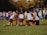 A group of young athletes wearing maroon shorts and white shirts gather on a grassy field, preparing for an event or competition. They appear to be in a huddle, perhaps discussing strategy or motivating each other. In the background, other participants and spectators are visible, some standing close to a barrier with blue banners. Trees with autumn foliage can be seen beyond the scene.