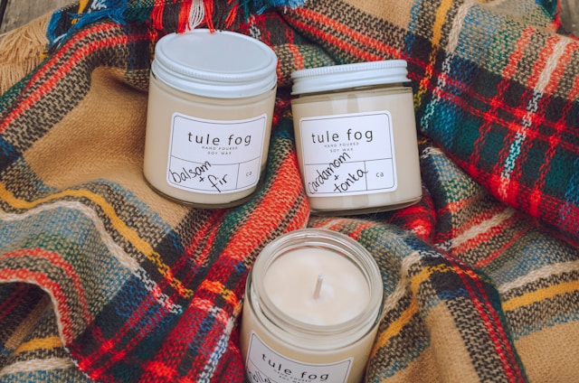 Three soy wax candles in glass jars labeled 'tule fog' are arranged on colorful plaid wool fabric. The candles are labeled with scents such as balsam and fir, and cardamom and tonka. The cozy and rustic setup suggests warmth and comfort.