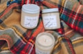 Three soy wax candles in glass jars labeled 'tule fog' are arranged on colorful plaid wool fabric. The candles are labeled with scents such as balsam and fir, and cardamom and tonka. The cozy and rustic setup suggests warmth and comfort.