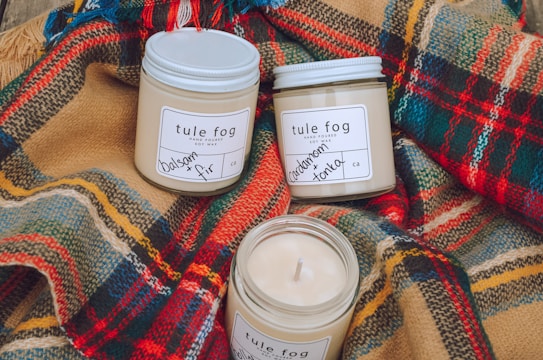 Three soy wax candles in glass jars labeled 'tule fog' are arranged on colorful plaid wool fabric. The candles are labeled with scents such as balsam and fir, and cardamom and tonka. The cozy and rustic setup suggests warmth and comfort.