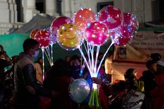 A group of people, including children, are gathered around a person holding a bouquet of illuminated balloons. The balloons are colorful, with some featuring smiley faces, and are lit from within, creating a vibrant and festive atmosphere. The scene appears to be in an outdoor market or fair, with a backdrop of tents and signage.