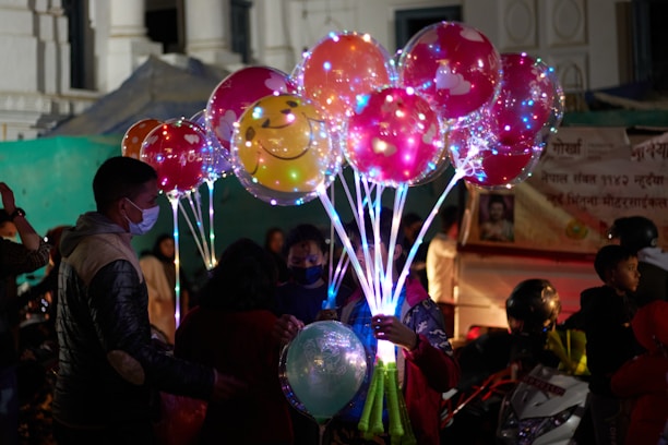 A group of people, including children, are gathered around a person holding a bouquet of illuminated balloons. The balloons are colorful, with some featuring smiley faces, and are lit from within, creating a vibrant and festive atmosphere. The scene appears to be in an outdoor market or fair, with a backdrop of tents and signage.
