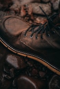 A rugged brown leather boot with lace-up details rests on a surface of wet, smooth stones. The setting suggests an outdoor environment, evoking a sense of adventure or exploration.