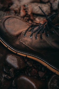 A rugged brown leather boot with lace-up details rests on a surface of wet, smooth stones. The setting suggests an outdoor environment, evoking a sense of adventure or exploration.