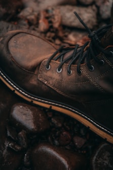 A rugged brown leather boot with lace-up details rests on a surface of wet, smooth stones. The setting suggests an outdoor environment, evoking a sense of adventure or exploration.
