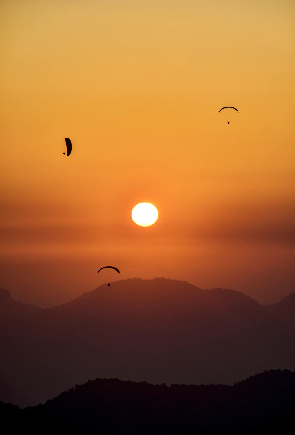 Three paragliders soar above a mountain range during a breathtaking sunset with vibrant oranges