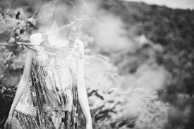 A black and white artistic photograph of a person wearing a patterned dress, standing amidst natural foliage. The image has a soft focus, giving it a dreamlike, ethereal quality. The background is blurred, creating a sense of depth and isolation.