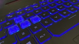 The image features a close-up view of a black keyboard with backlit blue LED lighting. Keys like W, A, S, and D are prominently visible, with the lighting highlighting their edges and letters.