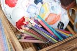 A wooden box holds a bundle of colorful pencils bound with a blue rubber band. There is a paint palette with red, blue, and yellow paints and a partially visible paintbrush nearby. The scene suggests a creative or artistic setting.