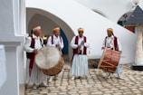 Four men in traditional attire are playing musical instruments. Two of them are holding large drums, one is playing a flute-like instrument, and the other is holding another drum. They are standing on a stone courtyard surrounded by white walls and arches.