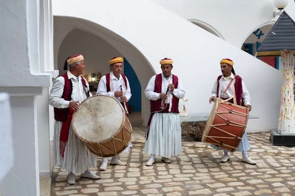 Four men in traditional attire are playing musical instruments. Two of them are holding large drums, one is playing a flute-like instrument, and the other is holding another drum. They are standing on a stone courtyard surrounded by white walls and arches.