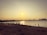 A beach scene at sunset with several people scattered along the shoreline. Some individuals appear to be taking photographs, including a couple dressed in wedding attire. The city skyline is visible across the water.