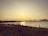 A beach scene at sunset with several people scattered along the shoreline. Some individuals appear to be taking photographs, including a couple dressed in wedding attire. The city skyline is visible across the water.