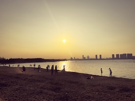 A beach scene at sunset with several people scattered along the shoreline. Some individuals appear to be taking photographs, including a couple dressed in wedding attire. The city skyline is visible across the water.