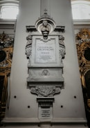 A detailed stone monument with a carved bust on top, intricate designs, and inscriptions in both Polish and English. It is set against a light-colored wall inside a building with other ornamental features visible in the background.