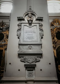 A detailed stone monument with a carved bust on top, intricate designs, and inscriptions in both Polish and English. It is set against a light-colored wall inside a building with other ornamental features visible in the background.