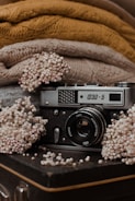 A vintage camera is surrounded by delicate, dried flowers on a textured, dark surface. Behind the camera, there are cozy, knitted fabrics in shades of brown and beige, adding a warm, rustic feel to the scene.