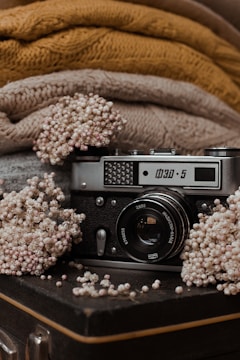 A vintage camera is surrounded by delicate, dried flowers on a textured, dark surface. Behind the camera, there are cozy, knitted fabrics in shades of brown and beige, adding a warm, rustic feel to the scene.