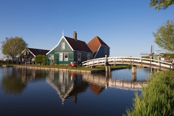 A quaint rural scene featuring a traditional green wooden house with a red-tiled roof, located beside a tranquil canal. A small white bridge arcs over the water, leading to the house. The surroundings include lush greenery and trees, with a classic Dutch windmill in the distance under a clear blue sky.