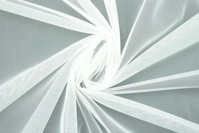 A close-up view of white, translucent fabric arranged in a swirling pattern. The intricate texture of the mesh is visible, creating a delicate and airy appearance.