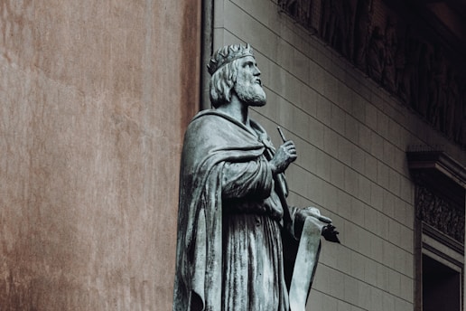 A stone statue of a historical figure wearing a crown and cape, holding a scroll and a pen, stands against the backdrop of a building with classical architectural details.