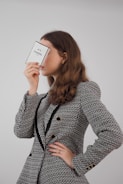 A person with long brown hair is holding a perfume box labeled 'N°5 Chanel PARIS' close to their face, obscuring it partially. They are dressed in a textured, patterned jacket with black accents and gold buttons.