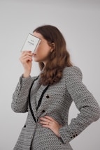 A person with long brown hair is holding a perfume box labeled 'N°5 Chanel PARIS' close to their face, obscuring it partially. They are dressed in a textured, patterned jacket with black accents and gold buttons.