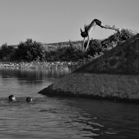 A person is captured mid-dive from a rocky ledge into a tranquil body of water, while two others are swimming nearby, creating an atmosphere of leisure and adventure. The surroundings are natural with lush vegetation on the banks.