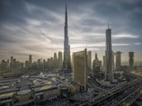 A cityscape with towering skyscrapers under a dramatic sky. The tallest building dominates the skyline, surrounded by other high-rise structures. A complex network of roads is visible, along with a large building featuring 'Dubai Mall' signage. The architecture is modern and sleek.