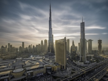 A cityscape with towering skyscrapers under a dramatic sky. The tallest building dominates the skyline, surrounded by other high-rise structures. A complex network of roads is visible, along with a large building featuring 'Dubai Mall' signage. The architecture is modern and sleek.