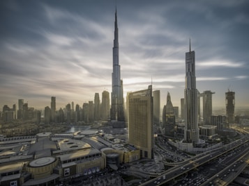 A cityscape with towering skyscrapers under a dramatic sky. The tallest building dominates the skyline, surrounded by other high-rise structures. A complex network of roads is visible, along with a large building featuring 'Dubai Mall' signage. The architecture is modern and sleek.