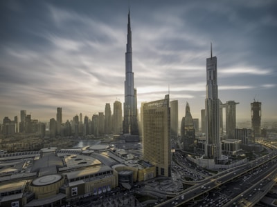 A cityscape with towering skyscrapers under a dramatic sky. The tallest building dominates the skyline, surrounded by other high-rise structures. A complex network of roads is visible, along with a large building featuring 'Dubai Mall' signage. The architecture is modern and sleek.