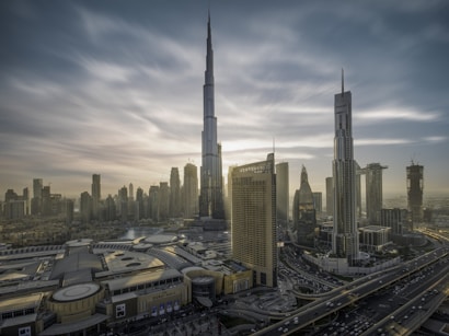 A cityscape with towering skyscrapers under a dramatic sky. The tallest building dominates the skyline, surrounded by other high-rise structures. A complex network of roads is visible, along with a large building featuring 'Dubai Mall' signage. The architecture is modern and sleek.