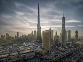 A cityscape with towering skyscrapers under a dramatic sky. The tallest building dominates the skyline, surrounded by other high-rise structures. A complex network of roads is visible, along with a large building featuring 'Dubai Mall' signage. The architecture is modern and sleek.